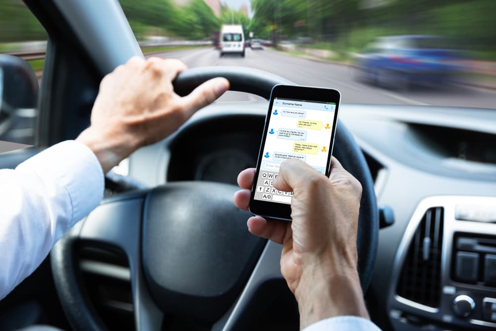 Close-up of a man's hand typing a text message on a mobile phone while driving a car, illustrating the danger of distracted driving.