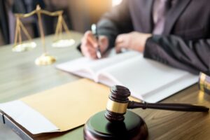 A gavel rests on a sound block on a wooden desk, symbolizing justice and law, as a lawyer works in the background.