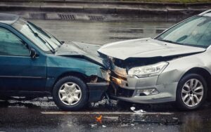 A rainy street scene showing the aftermath of a head-on collision between two cars. The vehicles are visibly damaged, with crumpled front ends. The wet road surface and overcast sky indicate ongoing rain, contributing to hazardous driving conditions. The image highlights the consequences of a car crash, possibly due to traffic rule violations.