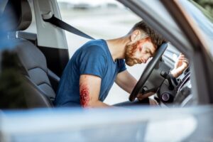 A close-up view inside a car shows a driver, still fastened with a seatbelt, appearing dazed and in shock after a road accident. The driver has deep bleeding injuries, with visible blood on their face or body. The interior of the car is slightly damaged, emphasizing the severity of the crash. The scene conveys distress and the urgent need for medical attention.