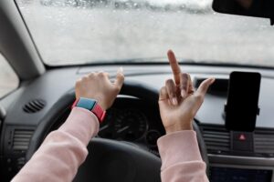 An upset millennial African American woman is driving a car, visibly frustrated, with her hand gripping the steering wheel. She is showing the middle finger through the window, expressing anger, possibly due to traffic problems or a violation of traffic rules. The window is covered in raindrops, and the city road is visible in the background, adding to the tense and chaotic atmosphere of the scene.