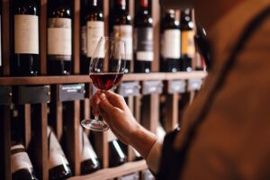 A male bartender or cavist stands near shelves stocked with wine bottles, holding a glass of wine in one hand. He examines the wine's color by looking at the tint through the glass, while also smelling its aroma. The scene highlights his expertise in wine tasting, with the shelves filled with various wine bottles in the background.