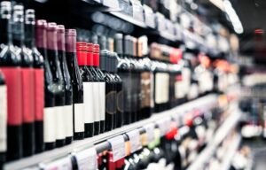 A shelf in a liquor store displays a selection of red wine bottles, including varieties like Pinot Noir, Merlot, and Cabernet Sauvignon. The bottles are neatly arranged, with the focus on their labels and colors. In the background, a supermarket aisle is visible, suggesting a well-stocked store offering a range of alcoholic beverages for sale. The image emphasizes the variety and selection available to customers.