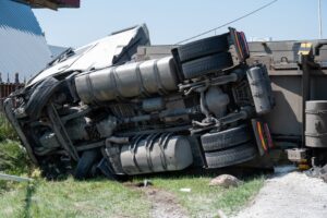 A large truck is overturned on its side in the middle of a highway following a car accident. Debris is scattered around the scene, and the damaged truck shows significant impact marks. The highway appears partially blocked, with other vehicles either stopped or navigating around the accident. The image highlights the severity of the crash and its impact on traffic flow.