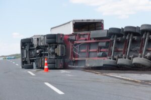 An overloaded truck (TIR) lies overturned on an interurban road after losing control. The truck’s cargo is visibly scattered around the scene, with debris across the road. The truck's side is heavily damaged, and the surrounding area shows signs of an accident, including skid marks. The image conveys the danger of overloaded vehicles and the impact of speeding on road safety.