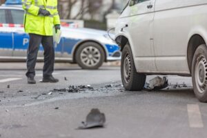 A police officer in uniform closely examines the damage to a van involved in an accident. The van shows visible dents and scratches, particularly on its front or side. The officer holds a notepad or device, appearing focused as they assess the scene. The background may include a road, other vehicles, or onlookers, emphasizing the context of a traffic incident.