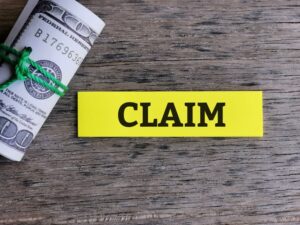 A sticky note with the word "CLAIM" written in bold letters is placed on top of a stack of fake money, resembling U.S. dollar bills. The scene symbolizes business and finance concepts, such as insurance claims, financial compensation, or reimbursement. The background is minimalistic, keeping the focus on the sticky note and money.