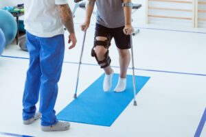 A man, wearing an orthosis on his leg and using crutches, is learning to walk in a clinic with the assistance of a supportive therapist standing nearby. The therapist is guiding him, offering encouragement and ensuring his safety as he takes cautious steps. The clinic environment is well-lit and equipped with rehabilitation tools, highlighting the process of recovery and physical therapy after a car accident.