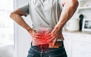 A man sits on the edge of a chair, visibly distressed, with one hand placed on his lower back, indicating pain. He leans slightly forward, grimacing in discomfort. The setting suggests a medical or home environment, with soft lighting emphasizing his struggle. The scene conveys the concept of lower back pain and the need for relief or medical intervention.