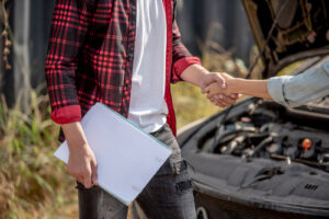 A person in a red plaid shirt holding documents shakes hands with another individual near a broken-down car with an open hood, symbolizing a post-accident discussion or insurance settlement.