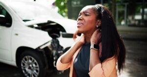 A woman standing in front of a damaged car holds her neck in pain, representing injuries and distress after a car accident.