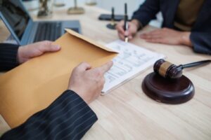 A lawyer hands over a legal document in an office setting with a gavel on the desk, symbolizing legal proceedings or settlement discussions.